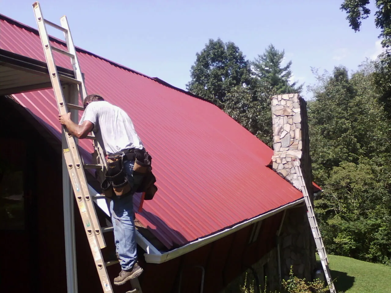 Roofer on a ladder beside a red metal roof and stone chimney — Soffit & Fascia Repair by Roofing Brothers in Dickinson